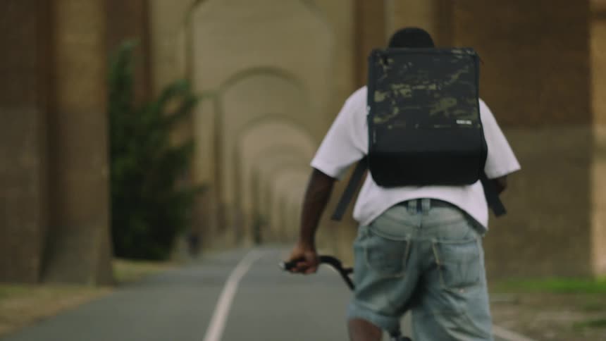 Male cyclist pedals along a highway passing under a series of building arches on a summer day
