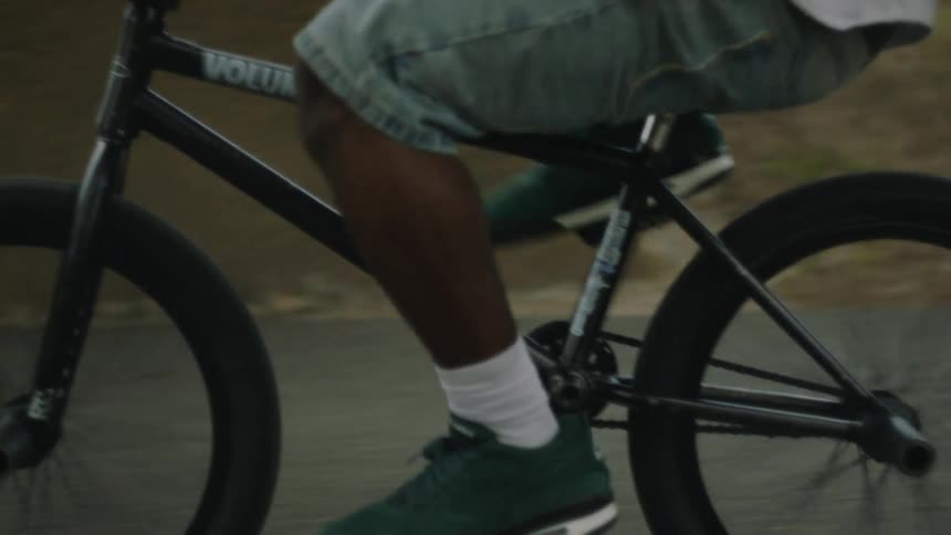 This is a closeup shot focused on the lower body of a person riding a black bmx bike on a paved road the rider is wearing denim shorts white crew socks and dark green athletic shoes as they pedal