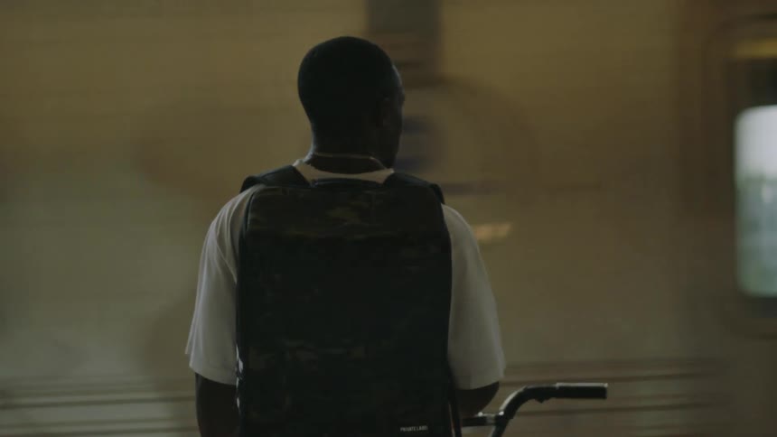 A rear view of a man wearing a white shirt a dark vest and a chain stands with a bicycle on an elevated train platform as a train rapidly passes obscuring the cityscape and bridge in the background