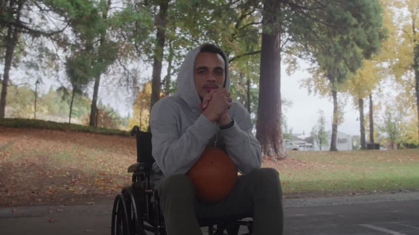 A man wearing a hooded sweatshirt sits in a black wheelchair holds an orange basketball on his lap and looks directly forward while positioned on a paved area surrounded by trees with autumn foliage