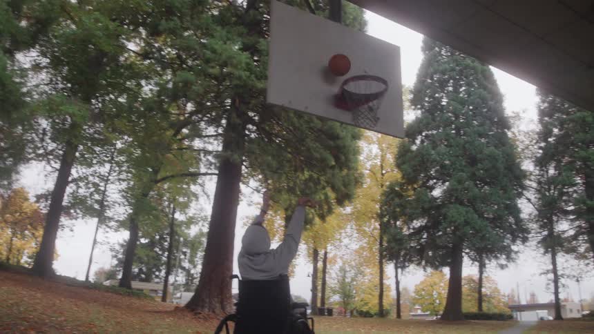 The video shows a person in a wheelchair moving away from the camera on an asphalt surface possibly a park basketball courtthe background is a treelined park with fall foliage creating a peaceful outdoor setting