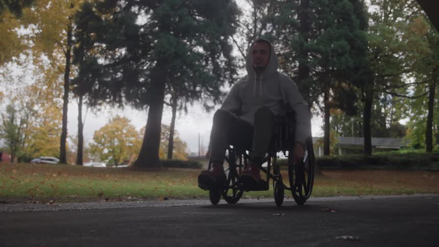 The video shows a man in a grey hoodie pushing himself forward in a manual wheelchair on an outdoor path he is smiling and moving along a paved path in a parklike setting with large trees in the background