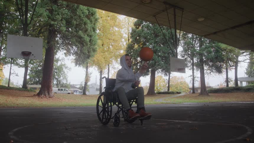 A young man in a wheelchair skillfully spins a basketball on his finger in the center of a covered outdoor basketball court surrounded by trees with autumn foliage