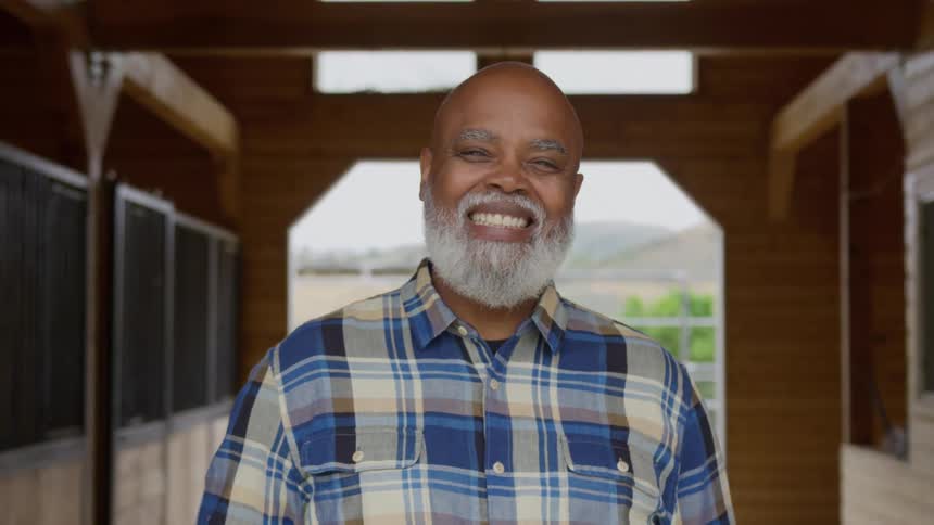 A close view shows a smiling bald bearded older man wearing a blue and brown plaid shirt standing inside a wooden barn or stable doorway