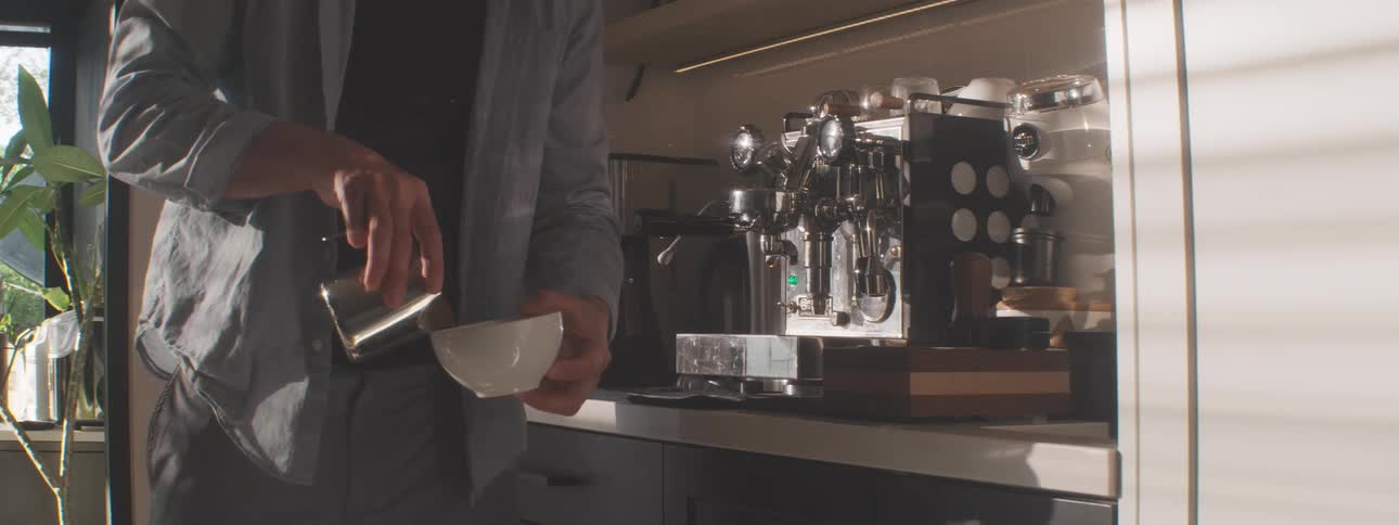 A person wearing a light shirt stands in a kitchen with a stainless steel coffee machine holds a metal pitcher with one hand and a white cup with the other and pours steamed liquid from the pitcher into the cup to complete a coffee drink
