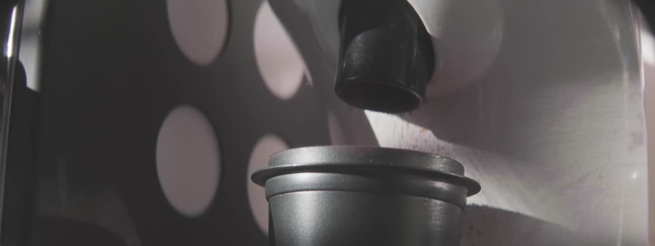 A close view of dark liquid dripping slowly from a black spout into a gray metal container with shadows and bright white circles visible on the wall in the background