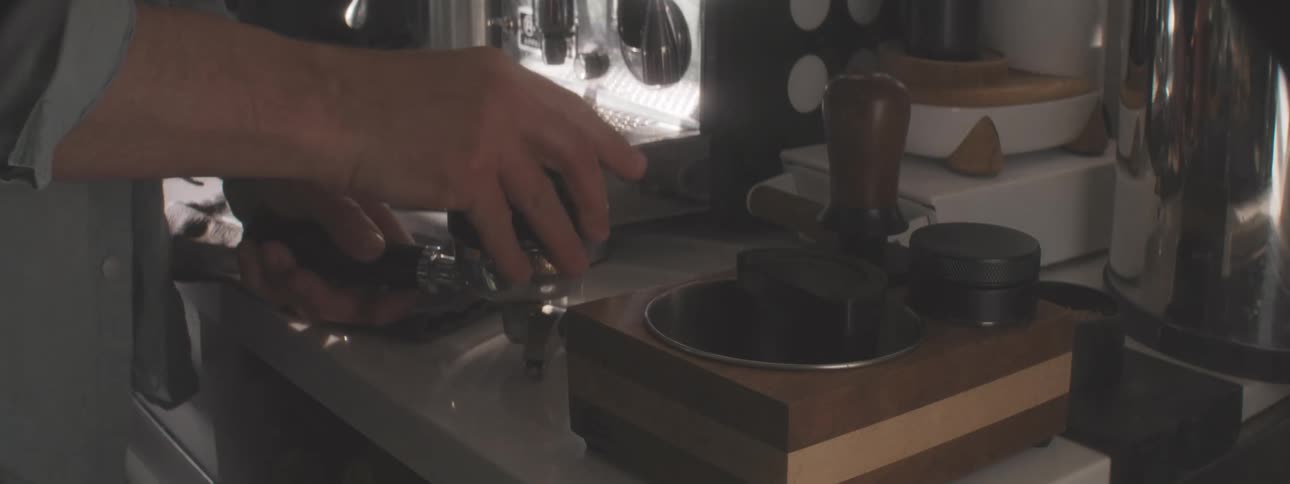 A persons hands hold a metal portafilter use a tamper to press down coffee grounds over a wooden block sweep excess grounds off the edge and wipe the area clean near a chrome espresso machine on a white countertop