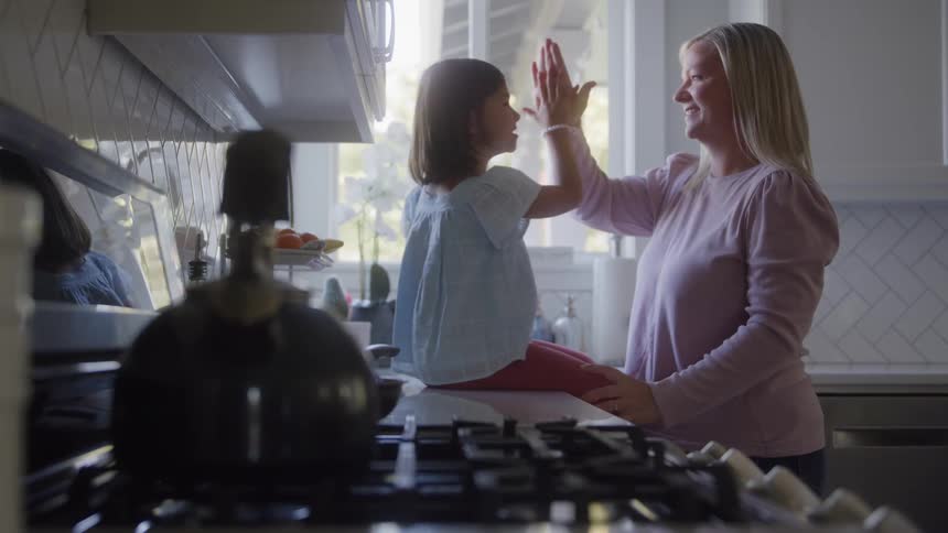 A woman stands in a bright white kitchen and talks with a girl sitting on the counter they exchange a palm slap gesture and a dark kettle and stovetop fill the immediate foreground