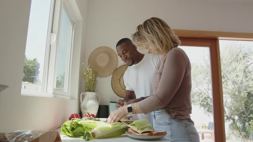 A man and a woman are smiling as they make sandwiches together