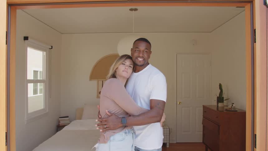 A smiling couple a man in a white shirt and a woman in a pink top and jeans stand embraced in the sunlit doorway of a bright bedroom with simple wooden furniture