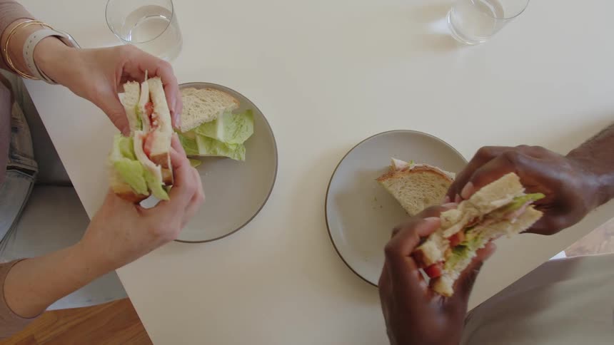 A topdown view shows two people eating sandwiches at a white table with one person wearing a smartwatch