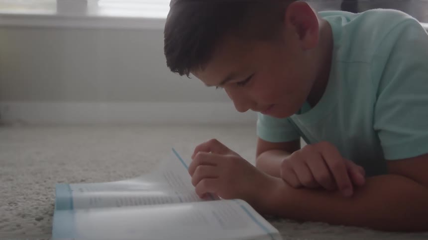 The video shows a young boy with dark hair and a light blue tshirt lying on a carpeted floor indoorshe is focused and intently reading an open book with his head resting on his forearms