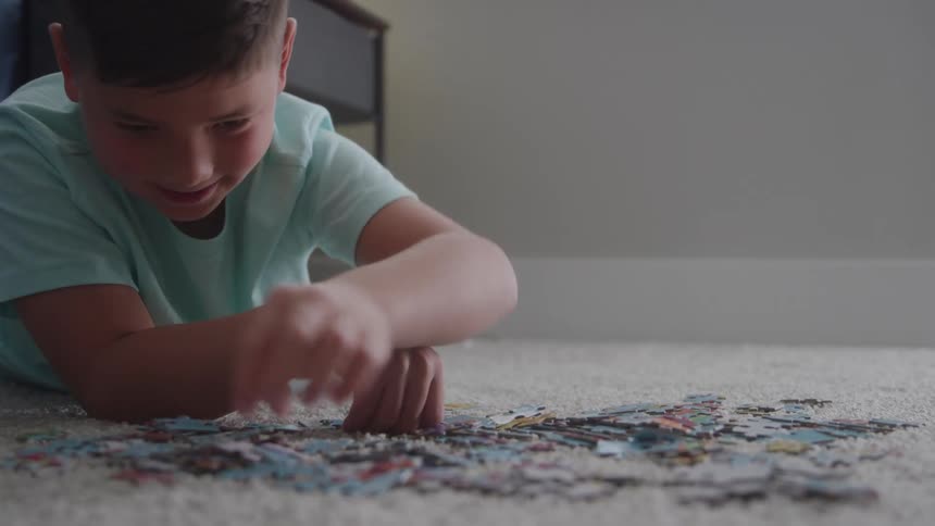 A young boy plays a jigsaw puzzle game on a rug in the middle of a room at midday