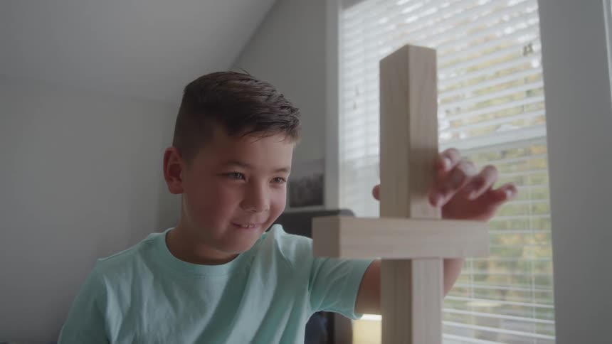 The video is a closeup of a young boy with dark hair and a light blue shirt smiling as he concentrates on an indoor activityhe is carefully stacking large wooden building blocks suggesting he is playing a game like jenga or building a tower