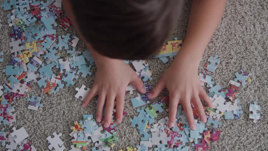 A child sits on the rug at midday sorting through scattered jigsaw puzzle pieces inside a room