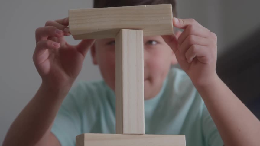 This is a closeup shot of a child playing with wooden blocks visible just above a vertical block the child carefully places a second block horizontally across the top of the first one to continue building a tower or structure