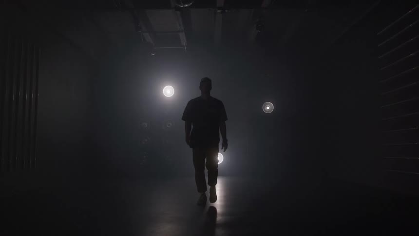 A person wearing dark clothes walks slowly from a distance toward the camera through a dark hazy room passing between several bright circular lights arranged on the back wall