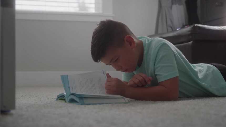 A young boy lays on his stomach on the carpet while reading a book