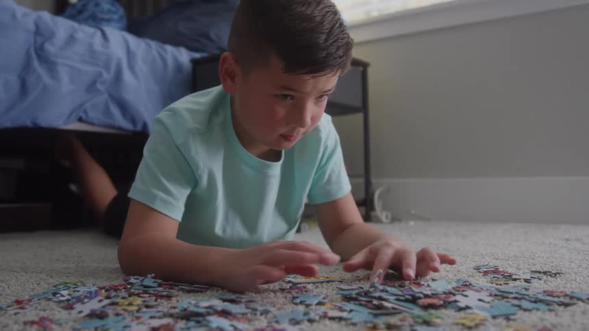 A young boy lies on the carpeted floor of his bedroom intently focused on sorting through a large pile of jigsaw puzzle pieces