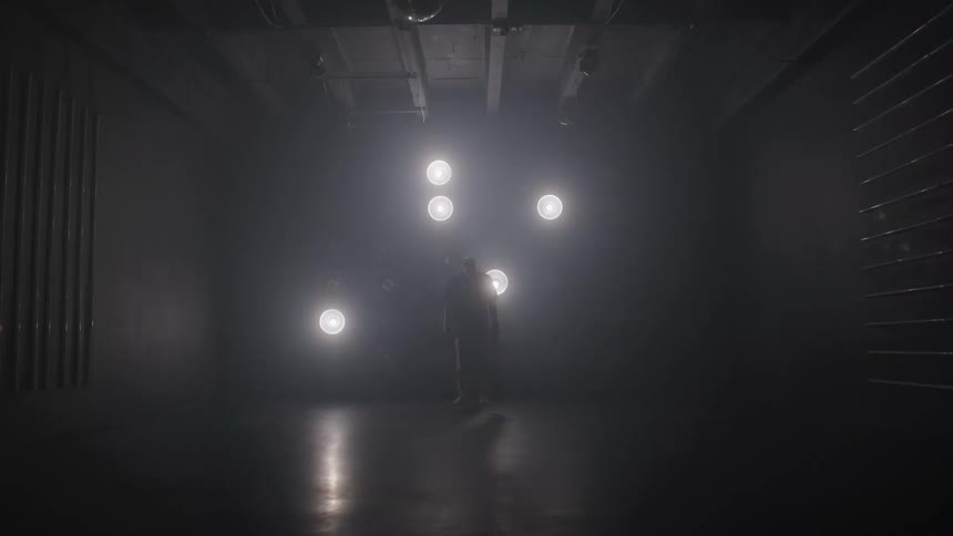 A dark video features a man standing in a large black room as several bright circular lights turn on and off in different patterns around him