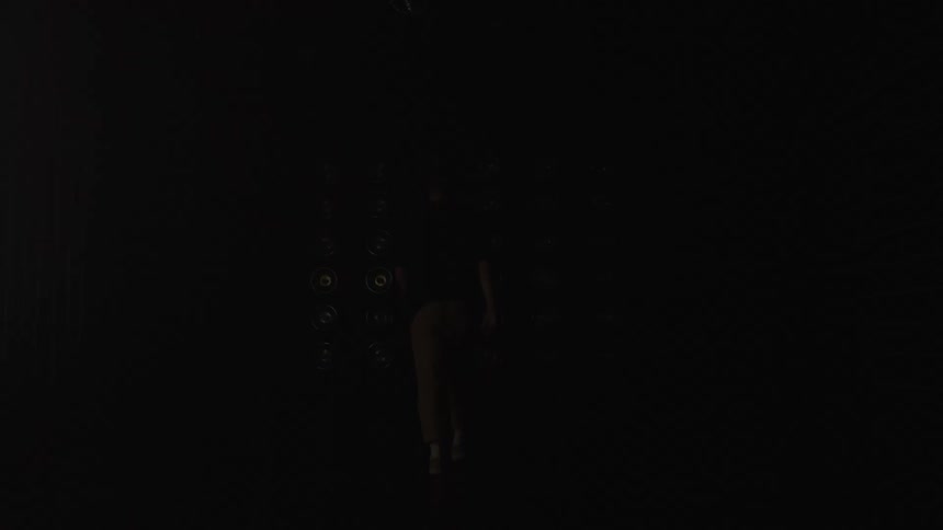 The video shows a person walking from a completely dark foreground toward a cluster of bright glowing circular lights set up on the floor of a large dark studio spacethe figure is seen only in silhouette against the illumination as they walk