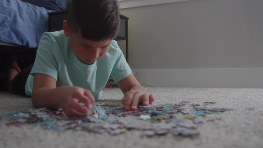 A young boy in a light blue shirt lies on a light gray carpet and concentrates on sorting the scattered pieces of a jigsaw puzzle in a bedroom