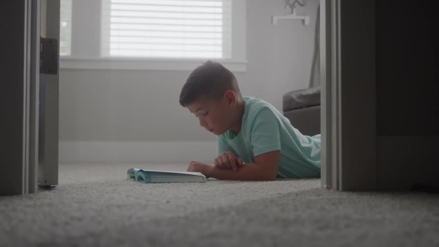 A young boy wearing a light blue shirt lies on a light gray carpet and reads a book visible through a doorway with bright window light in the background