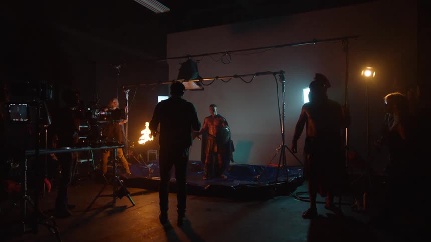 A film set is filled with crew members and camera equipment lights and a stunt performer with a flaming prop in a dark studio while an actor holds a sword in front of a white screen