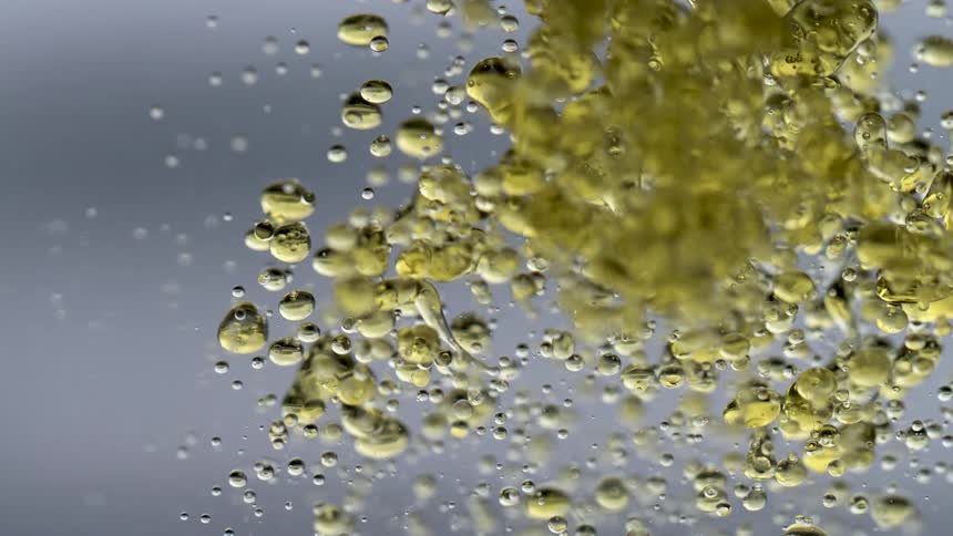 A closeup of golden liquid droplets and tiny air bubbles suspended in water against a soft gray background