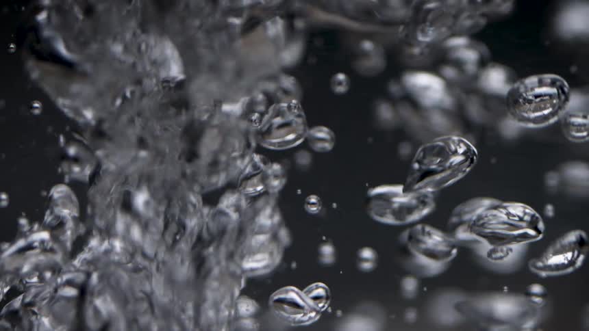 This is an extreme slowmotion closeup of water being forcefully poured or bubbling up against a dark background a multitude of smooth reflective air bubbles detach and rapidly rise past a turbulent column of water