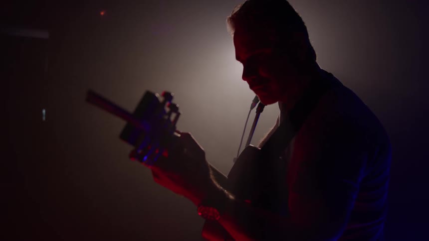 A close side view shows the silhouette of an older man with white hair smiling as he plays a dark electric guitar on a stage with red and blue ambient light