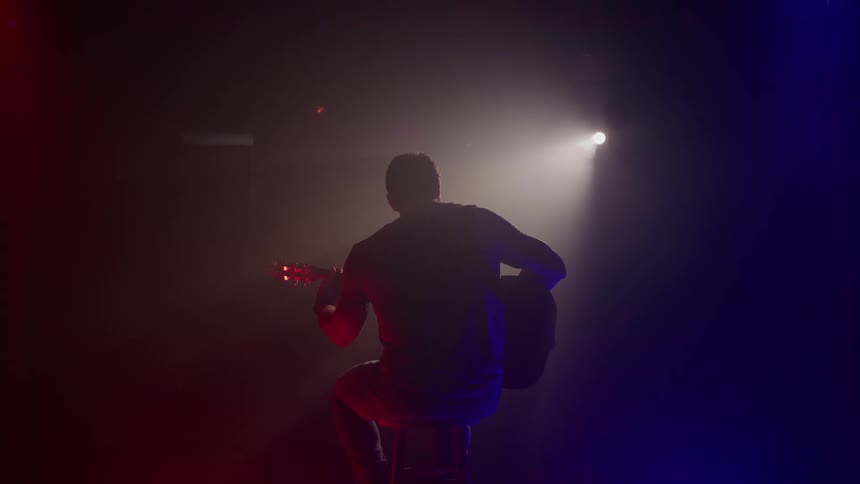 The man is sitting on a stool playing guitar