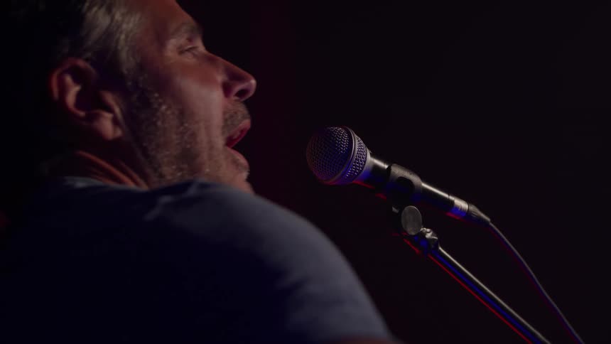 A close side view from behind shows an older man with a beard and a dark shirt leaning in to sing into a microphone on a stand with a subtle red glow in the dark background