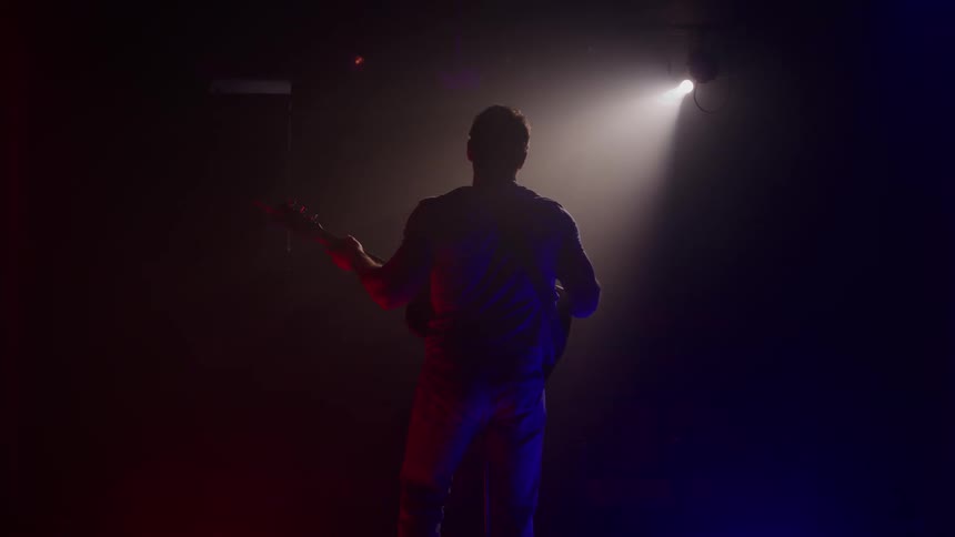 A silhouetted man stands on a stage with an electric guitar raising his hand toward a bright spotlight shining from above in a dark room illuminated by red and blue stage lighting