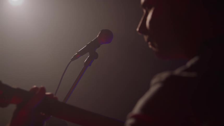A man wit ha guitar performs on a stage with his profile visible near a microphone the scene backlit by a single overhead light creating a strong silhouette against the dark background