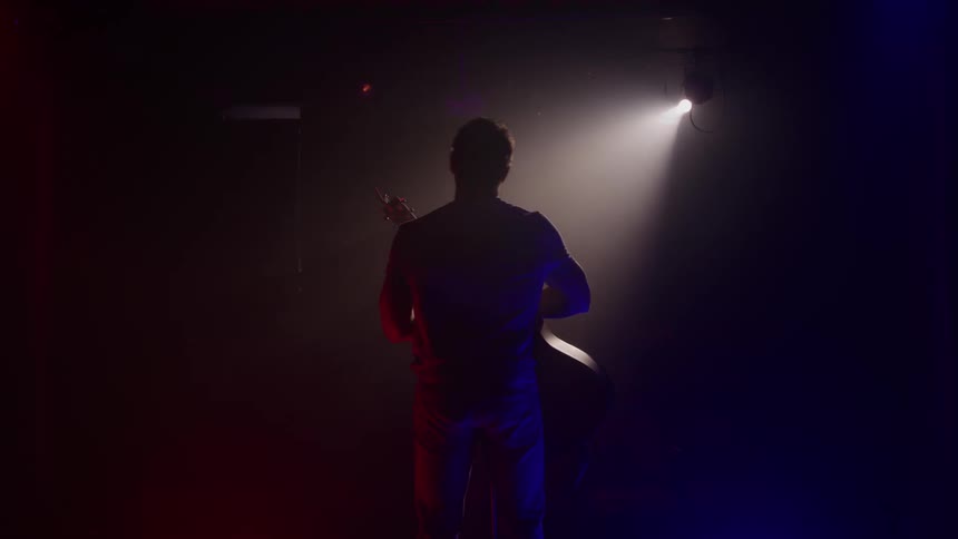 Male guitarist waves and bows on a dark stage lit by a spotlight