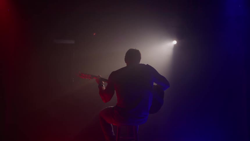 A musician sits facing away from the camera playing an acoustic guitar while silhouetted by a bright white spotlight and colored red and blue stage lighting on a dark stage