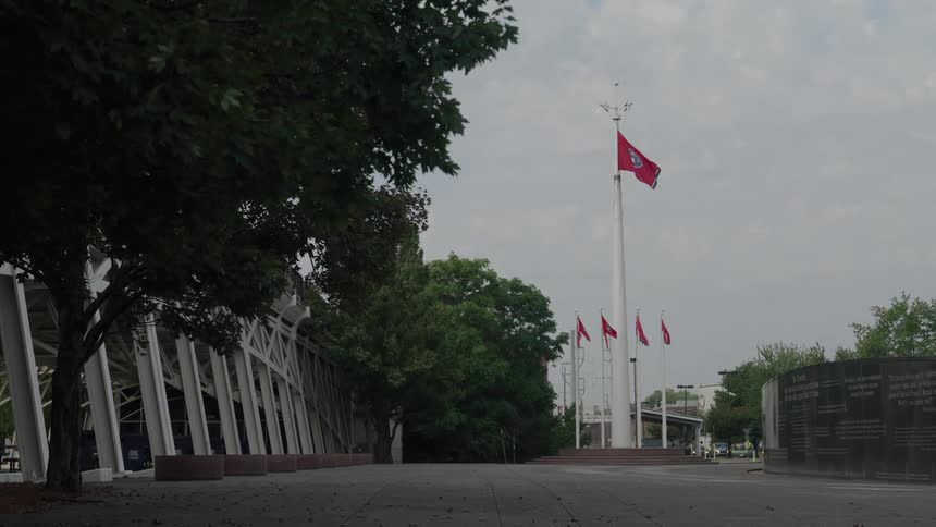 This is a static wide shot of an outdoor plaza or park entrance dominated by a tall flagpole flying the flag of tennessee a row of smaller red flags is visible below it with a large modern stadium structure to the left