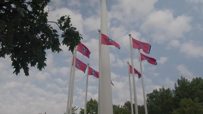 The video shows an establishing shot of the tennessee state capitol building and a modern skyscraper towering over a park with an outdoor amphitheaterin the foreground a tall central flagpole is surrounded by six smaller poles each flying a