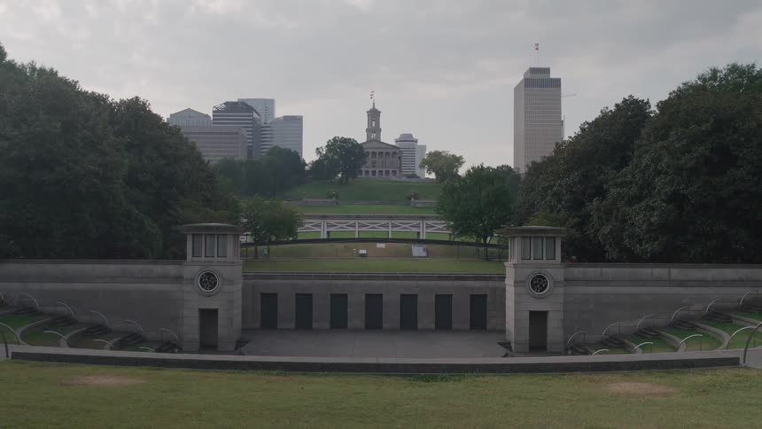 The video shows a wide shot of an outdoor amphitheater or grassy stadium with concrete structures and steps in the foregroundbeyond the green space and trees a city skyline with a domed building likely the tennessee state capitol and tall sk