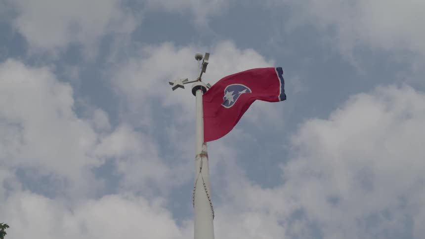 A low angle view shows the red flag of tennessee waving in the wind atop a white flagpole with a weather vane against a partially cloudy blue sky