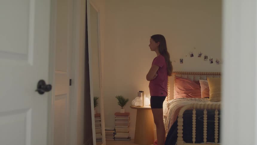 Young woman stands in front of a mirror in a warmly lit bedroom