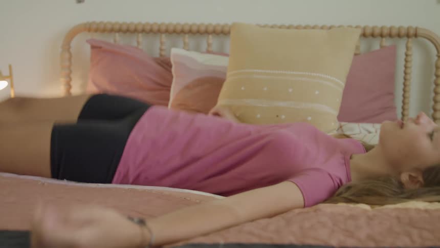A girl wearing a pink shirt and black shorts runs into a brightly lit bedroom and dramatically falls backward onto a bed with a wooden frame and decorative pillows