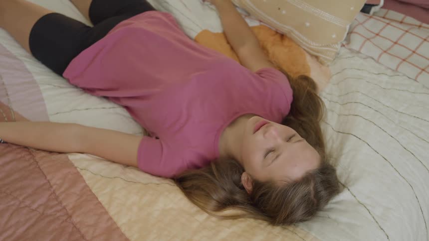 A teenage girl wearing a pink shirt and dark shorts lies supine with arms spread on a bed covered with a lightcolored patterned quilt resting motionless with eyes closed