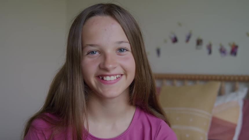 A young girl with long brown hair and blue eyes looks at the camera with a neutral expression before breaking into a wide happy smile in her bedroom