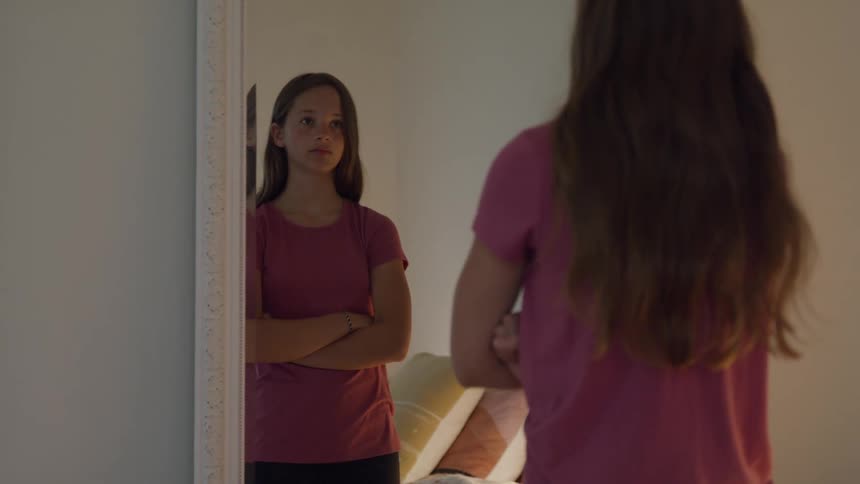 A girl in a pink shirt stands with folded arms looking at herself in a bedroom mirror appearing sad