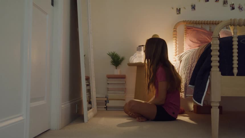 A long haired girl wearing a pink shirt and black shorts sits on the floor of her brightly lit bedroom looking in a full length mirror and adjusting her hair