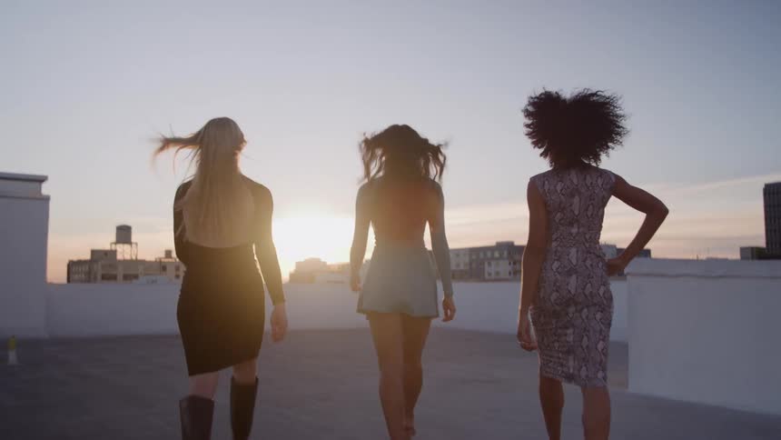 Three female models wearing fashionable dresses walk across a rooftop towards the sun as it sits on the horizon