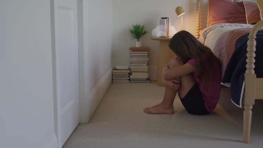 A teen girl with long hair is sitting on the bedroom floor and hugging her knees