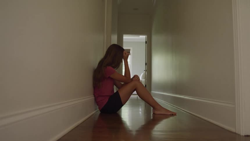 A sad long haired girl wearing a pink shirt and black shorts sits on the hardwood floor of a long dark hallway with her knees pulled up and her head resting on her hand